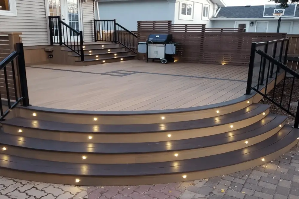 A beautiful custom-built deck by Dagmar Construction. Light-brown composite deck material is used to build rounded steps up to a deck containing furniture and a barbeque grill.