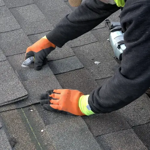 A contractor repairing shingles on a roof, wearing safety gloves and using a utility knife. Keep your roof in top shape with reliable repairs from Dagmar Pros LLC